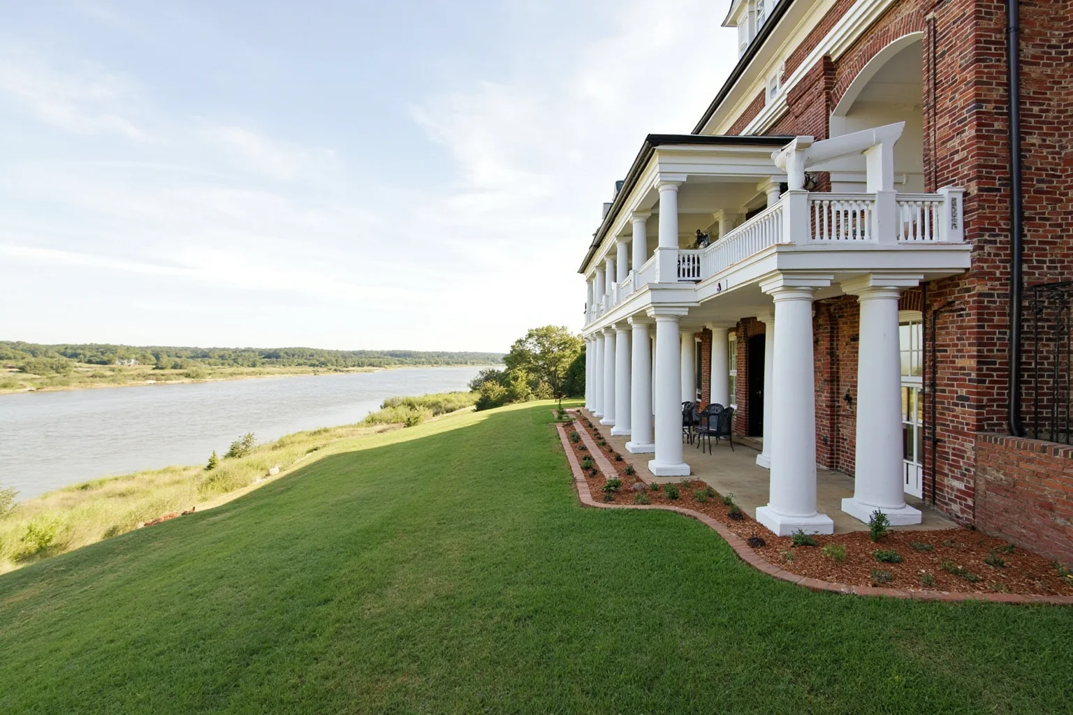 25 Famous American Historical Homes You Can Still Visit Today 2 Exterior view of a Georgian-style red-brick plantation mansion overlooking a wide river, with white columned porch and manicured lawn