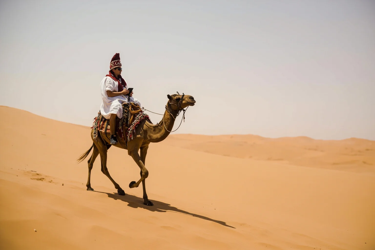 A British officer in desert robes riding a camel across a vast sand dune landscape