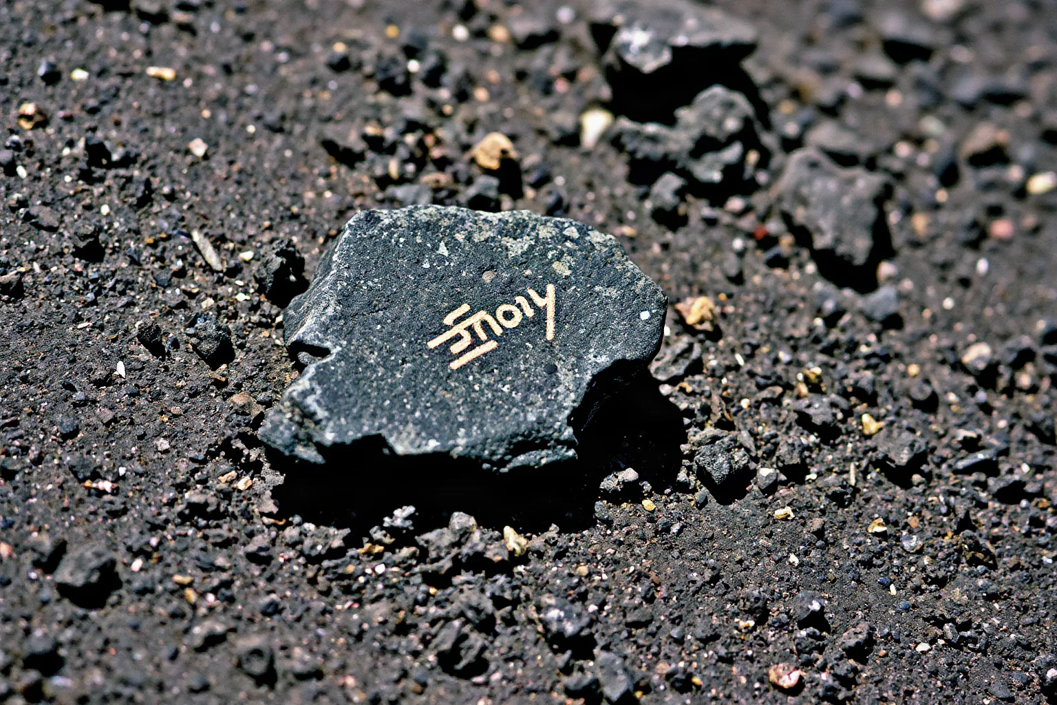 A close-up illustration of a small grey rock with a faint mark resembling a letter, lying on lunar soil