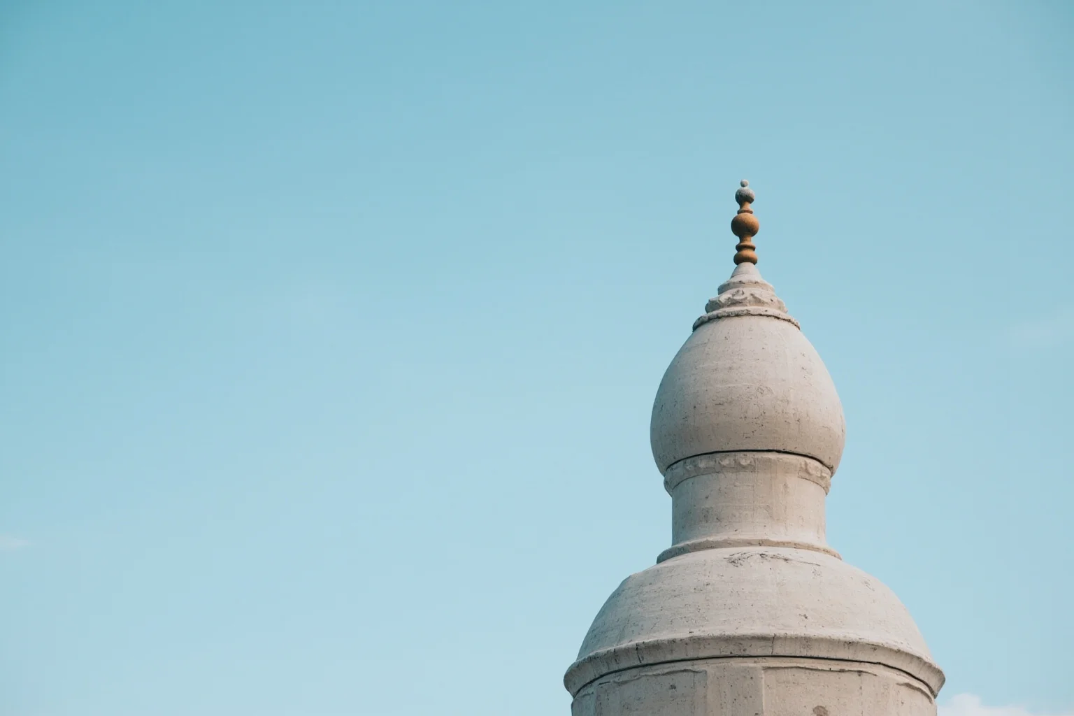 A close-up of a large bulbous white marble dome topped with an ornamental finial against a clear sky