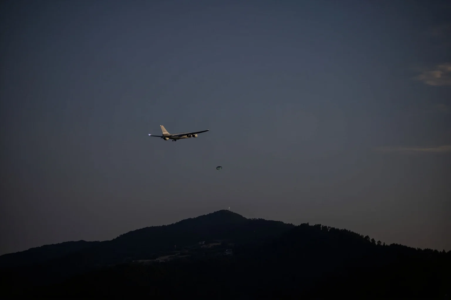 A commercial airplane at night over forested mountains with a parachute silhouette in the sky