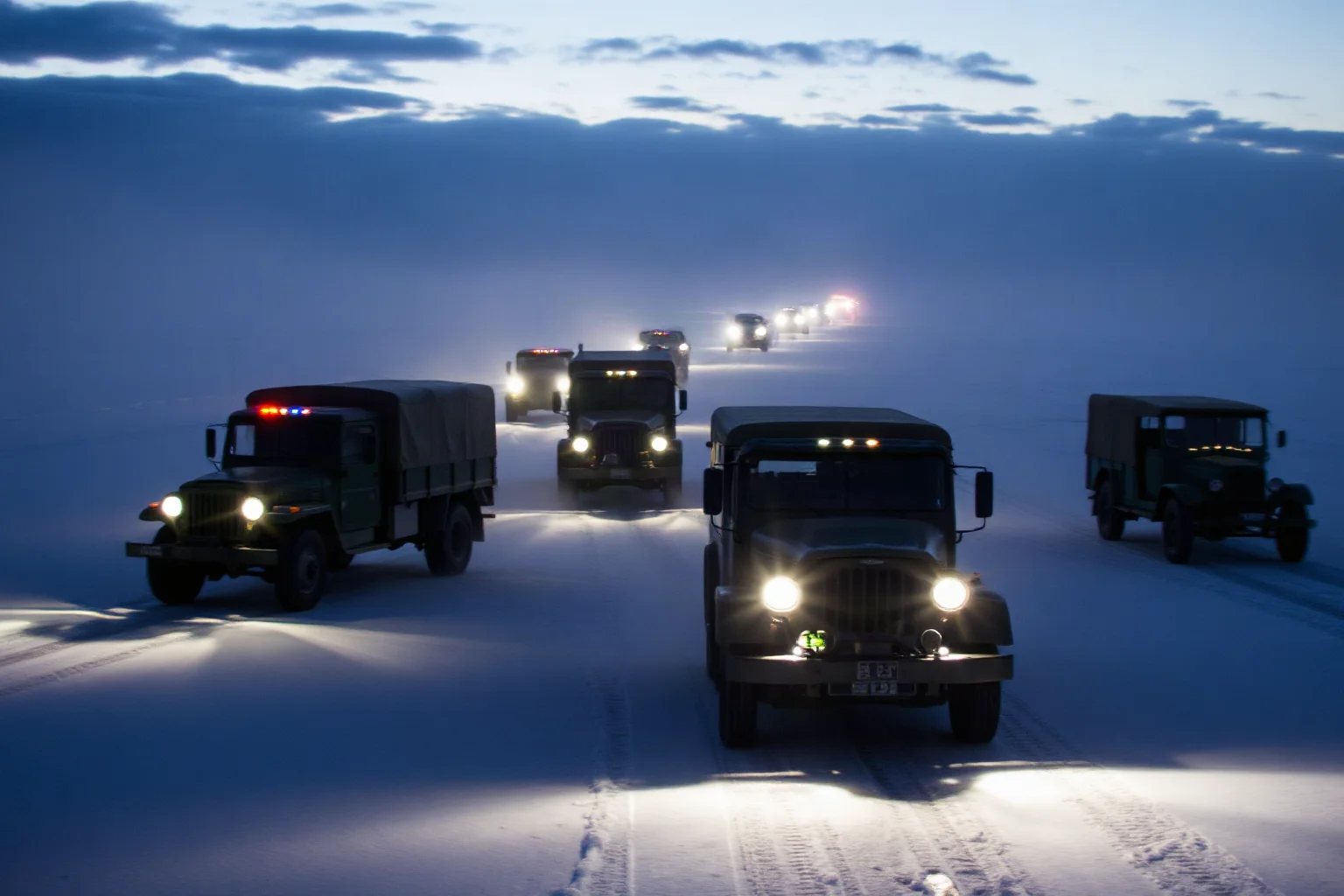 A convoy of old military trucks driving across a frozen lake at night under searchlights, ice and snow all around