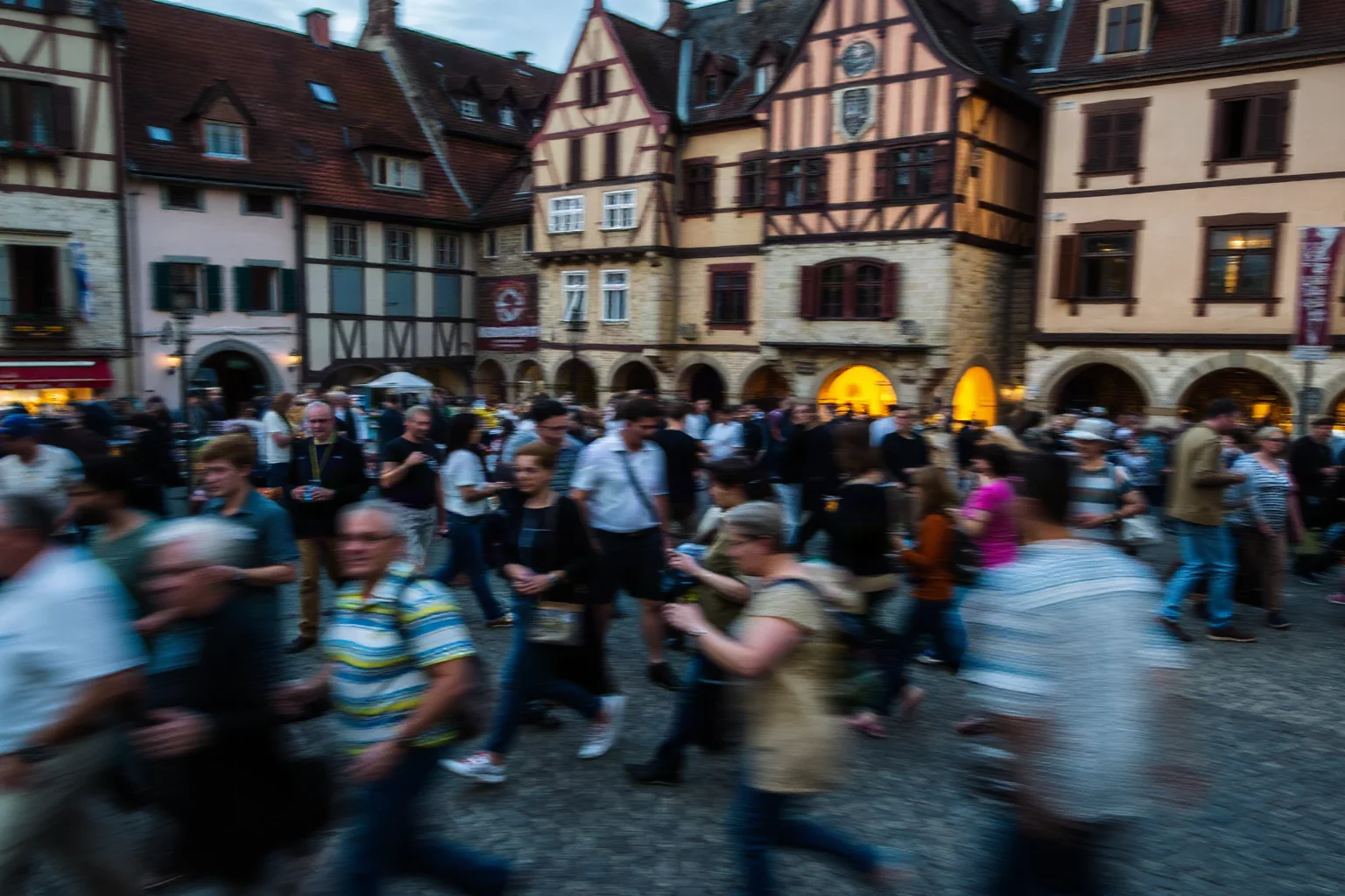 A crowded medieval European town square with blurred figures mid-motion, dusk lighting