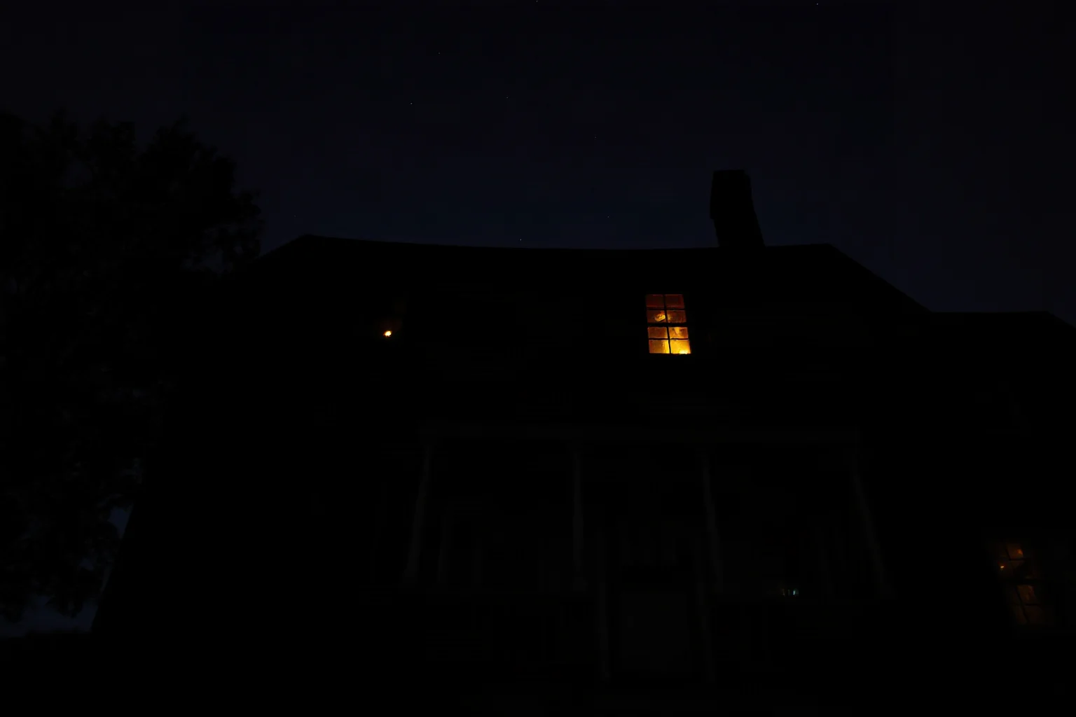 A dark 19th-century farmhouse at night with a faint eerie glow in an upstairs window, rural American gothic atmosphere