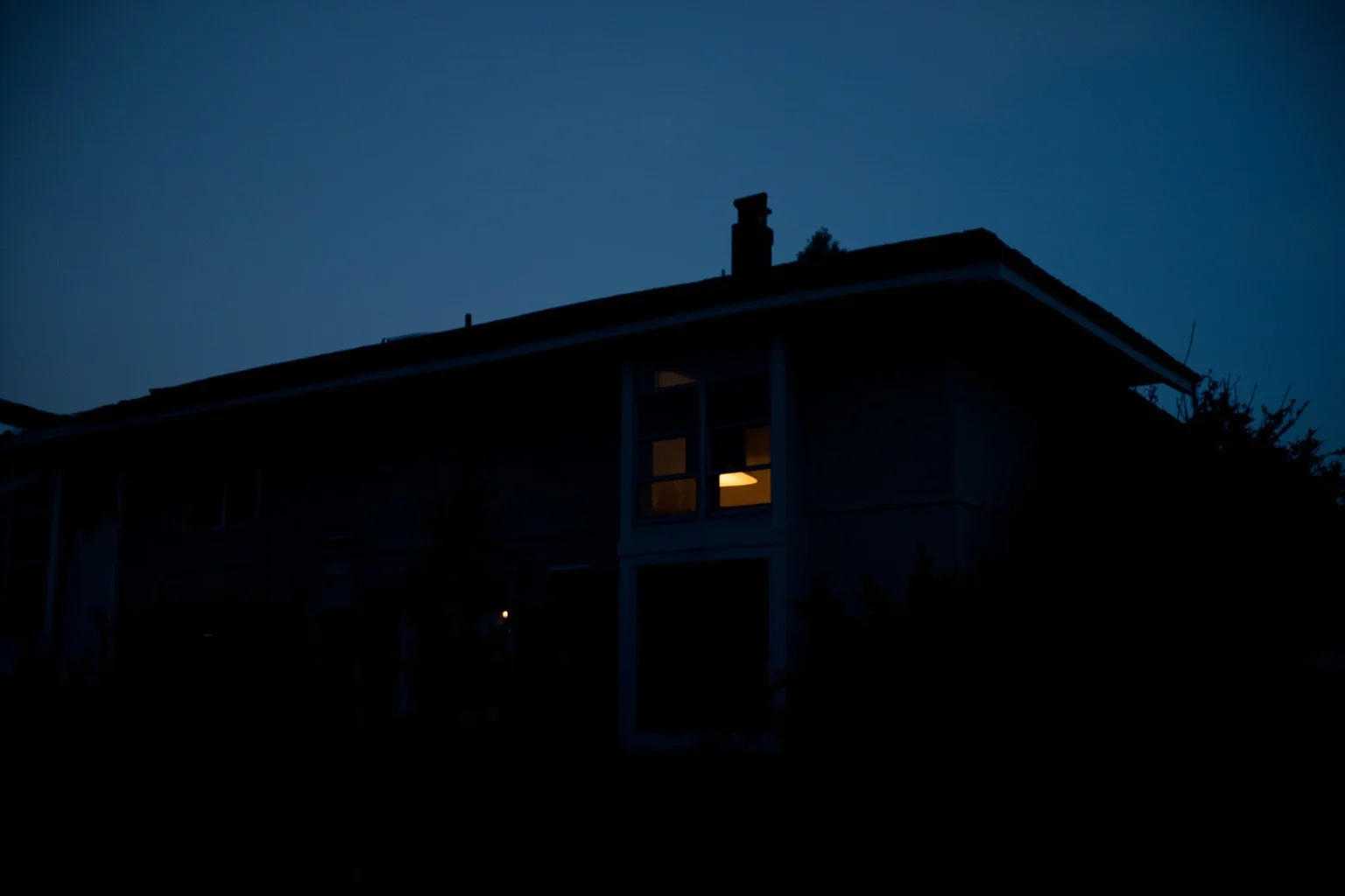 A dim suburban house at dusk with a single lit upstairs window