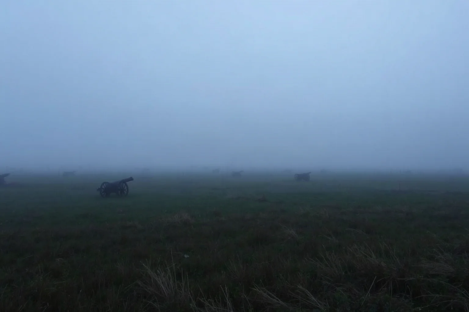 A foggy battlefield landscape with scattered fence lines and cannons at dawn, muted blue-grey tones, respectful non-graphic historical illustration.