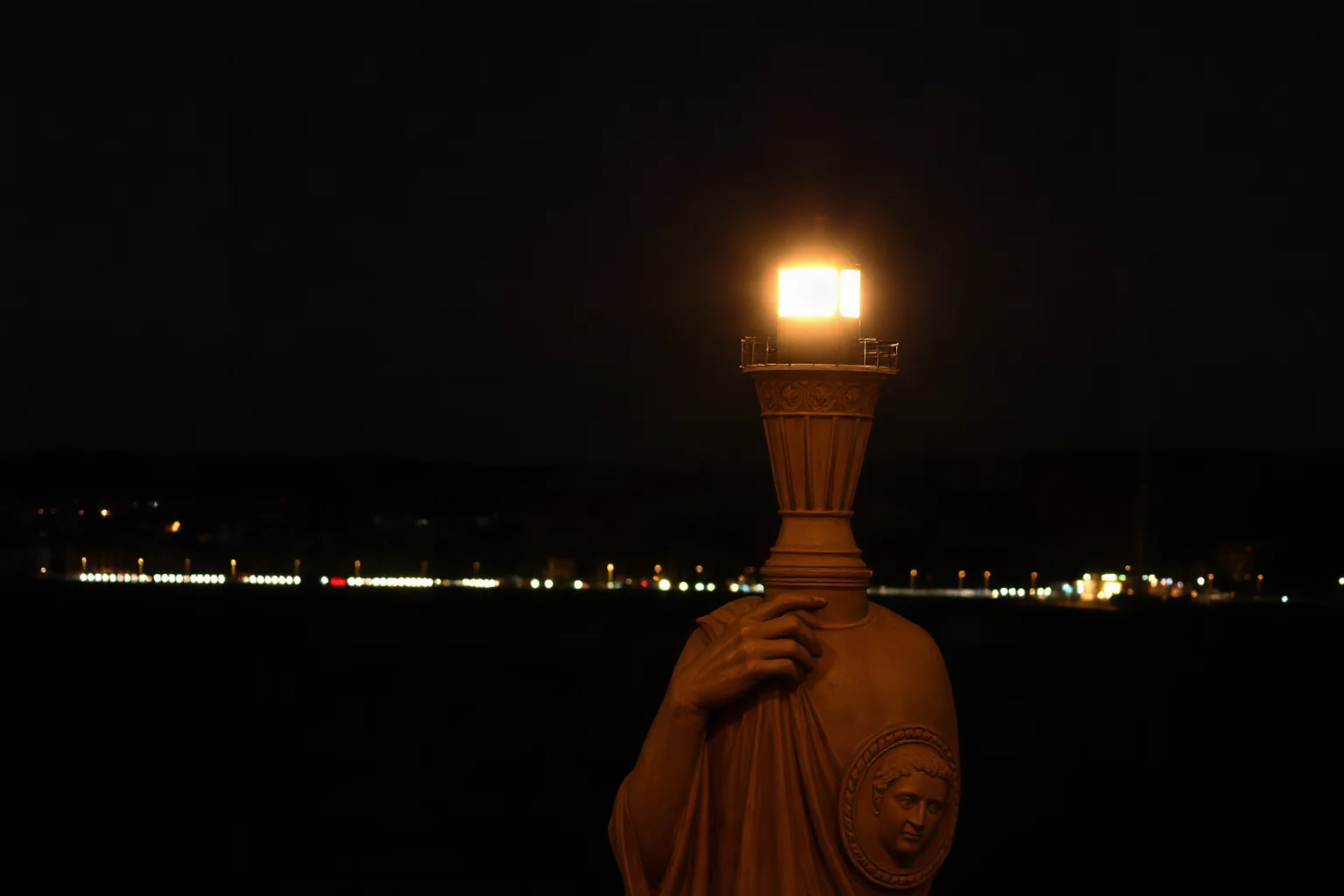 A glowing lighthouse beacon atop a statue's torch shining across a dark harbor at night