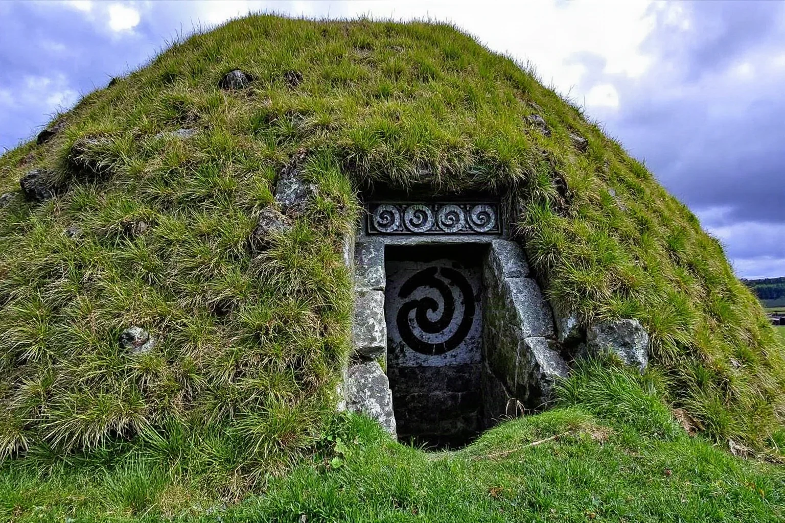 A large grass-covered passage tomb mound with a decorated stone entrance and carved spiral patterns