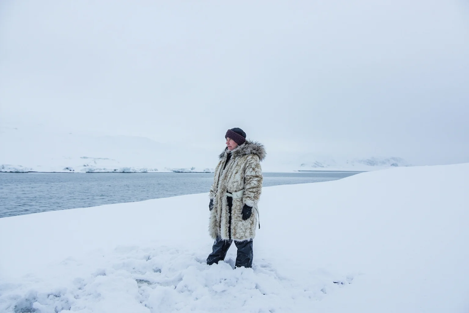 A lone figure in fur-lined Arctic clothing standing on a snowy remote island shoreline