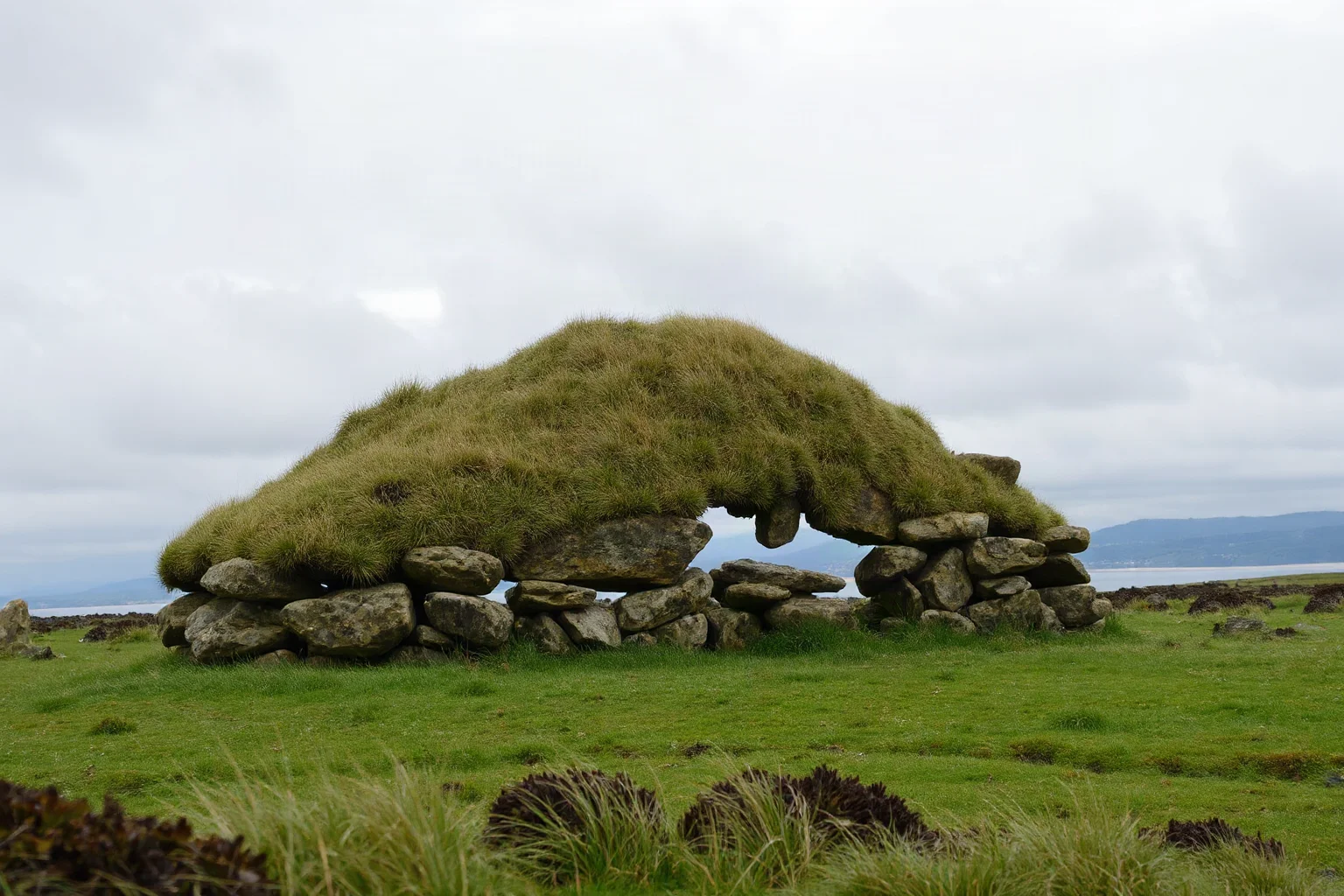 A massive elongated stone burial cairn covered in grass on a coastal European landscape under a cloudy sky