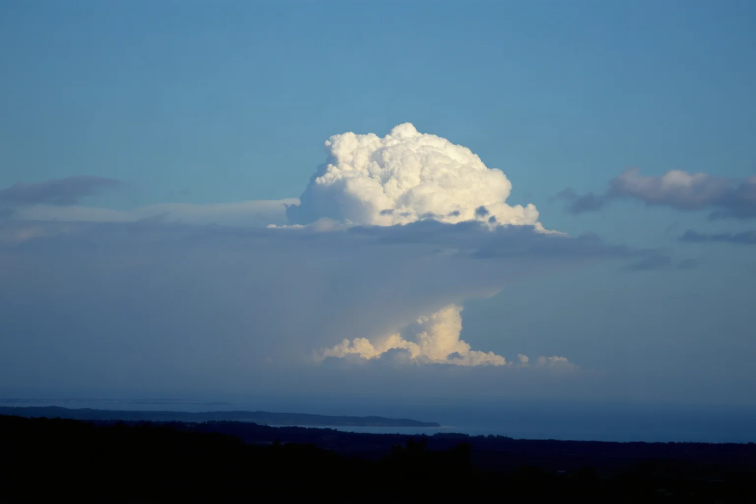 A mushroom cloud rising over a distant coastline, viewed from a safe silhouette vantage point