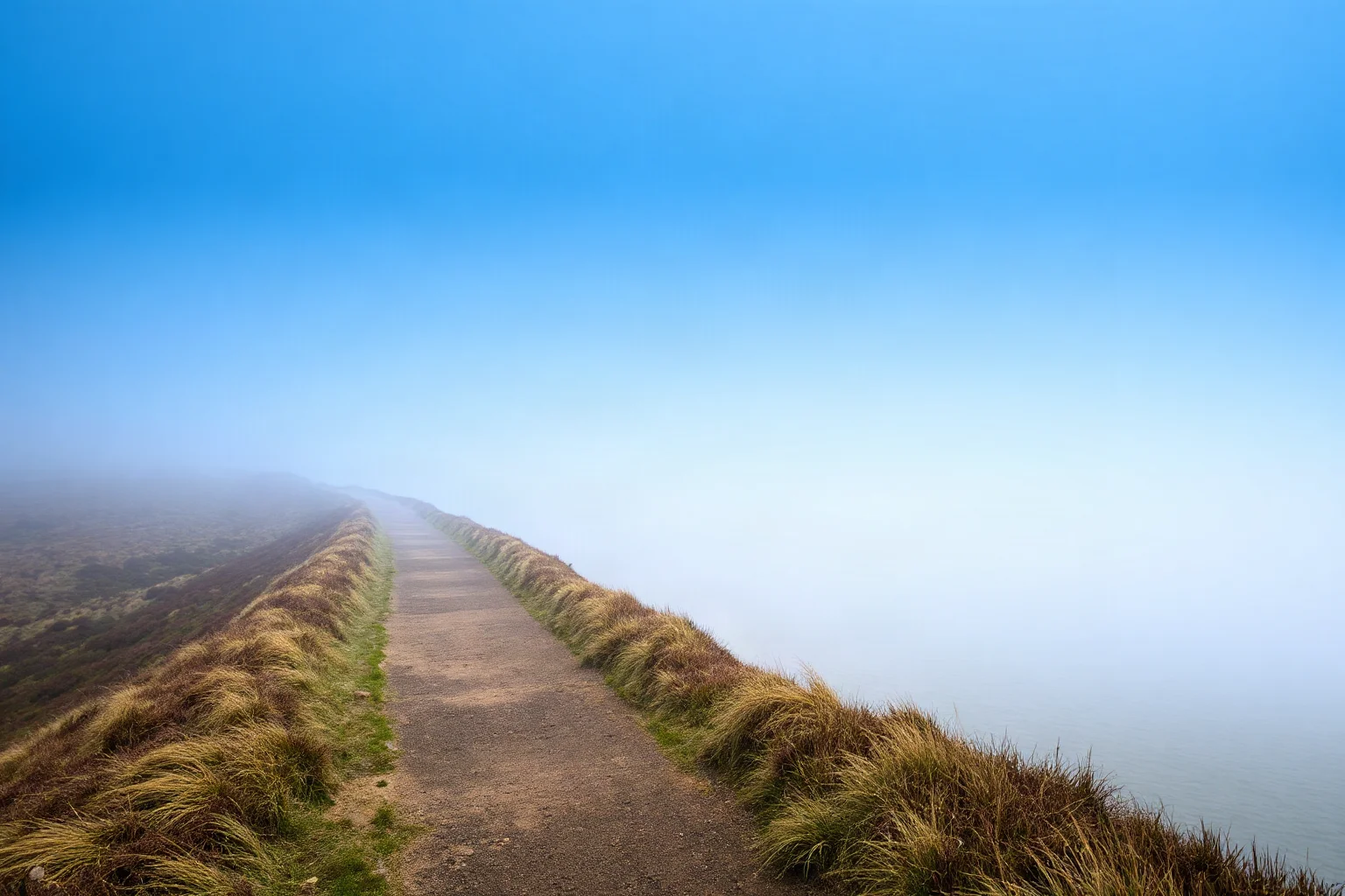 A narrow footpath leading toward a foggy coastline, symbolizing a long journey toward escape