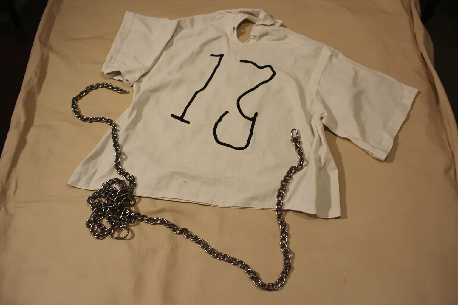 A numbered smock and ankle chain laid out on a bare cot, representing dehumanizing prisoner attire