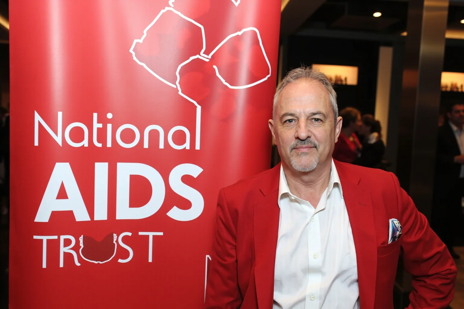 A patron figure standing beside a National AIDS Trust banner at a formal charity event