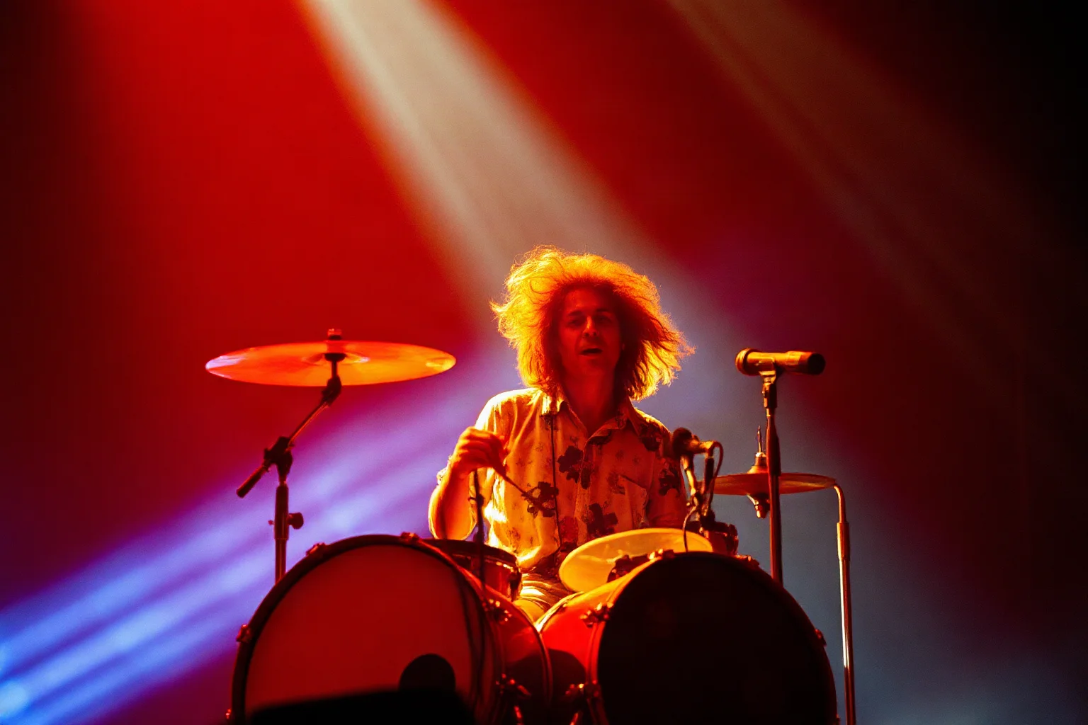 A psychedelic-era drummer with flowing hair performing energetically beside swirling stage light effects