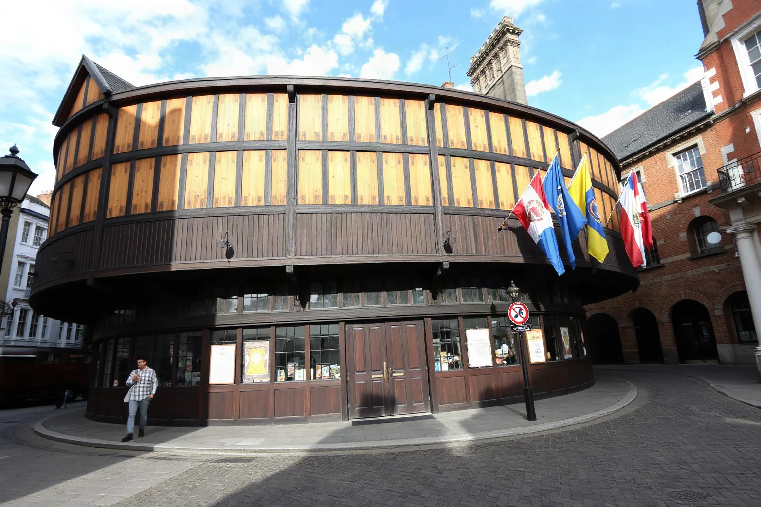A round wooden Elizabethan theater exterior (Globe-style) with flags, viewed from a cobblestone London street