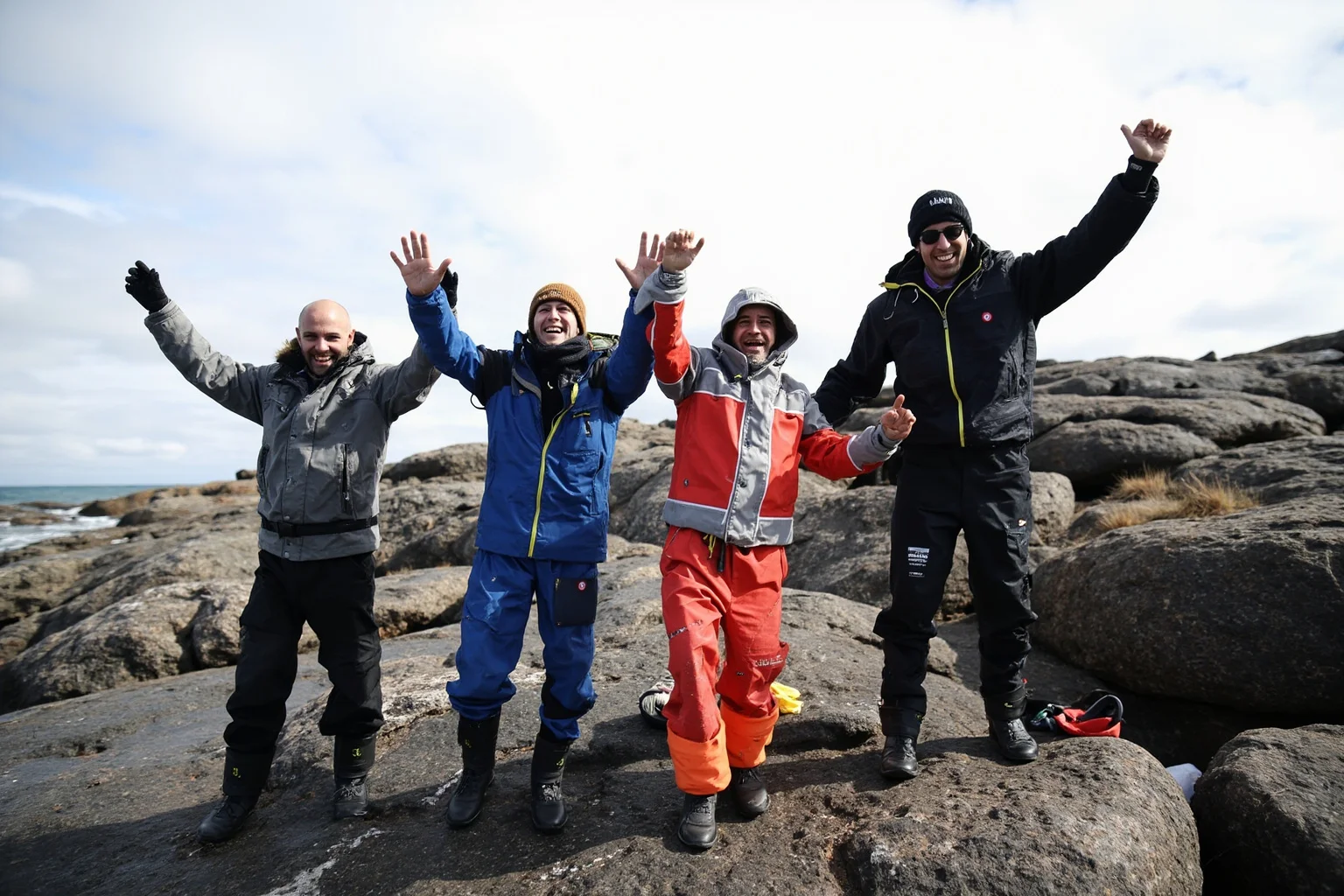 A small group of explorers in heavy expedition clothing celebrating survival on a rocky sub-Antarctic island shoreline