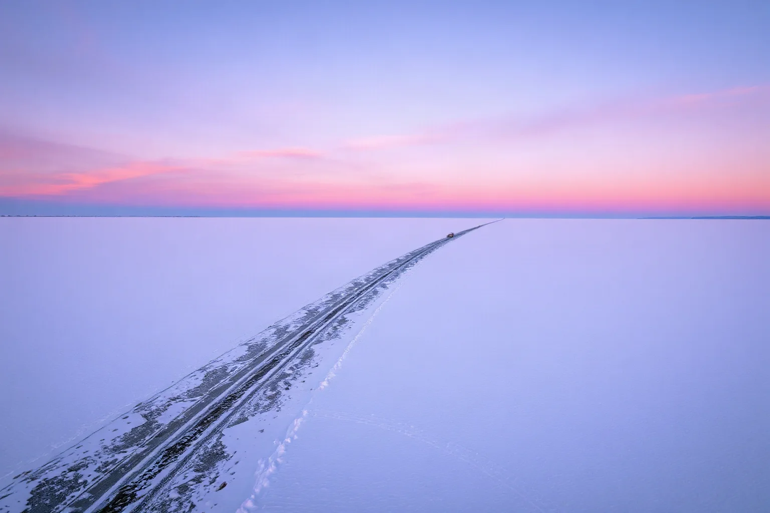 A snowy frozen lake at dusk with a single narrow supply route marked by faint truck tracks, isolated and vast