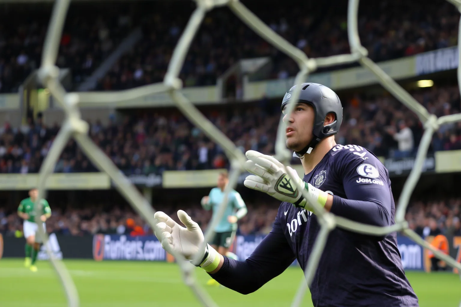 A soccer goalkeeper wearing a protective padded headguard making a save during a professional match, stadium atmosphere