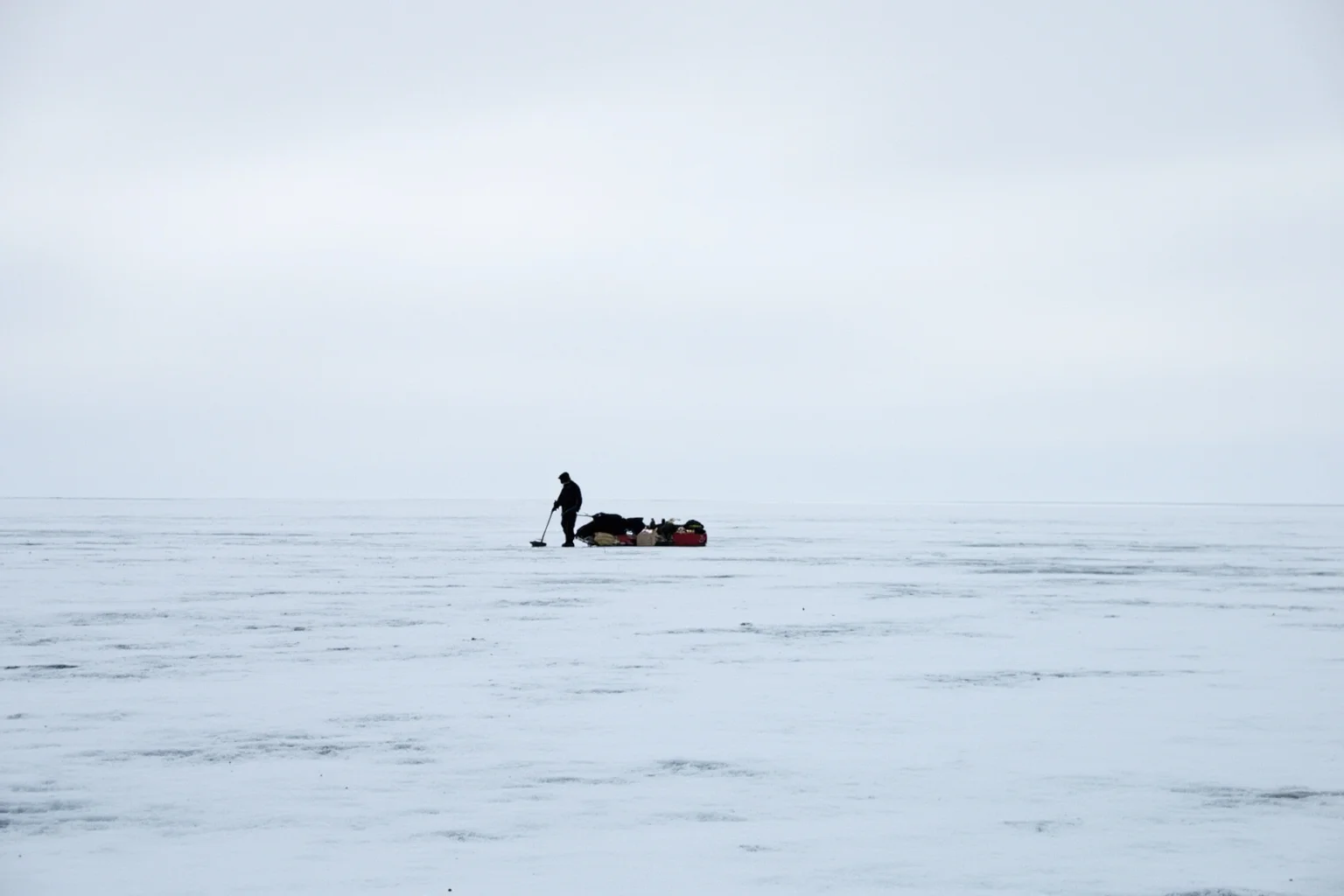 A solitary figure hauling a sledge across a vast icy Antarctic plateau under grey skies