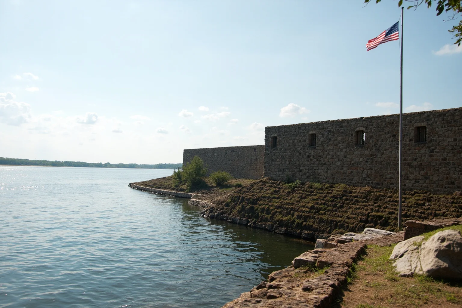 A stone-walled colonial-era fort beside a wide river with a flag flying above the ramparts