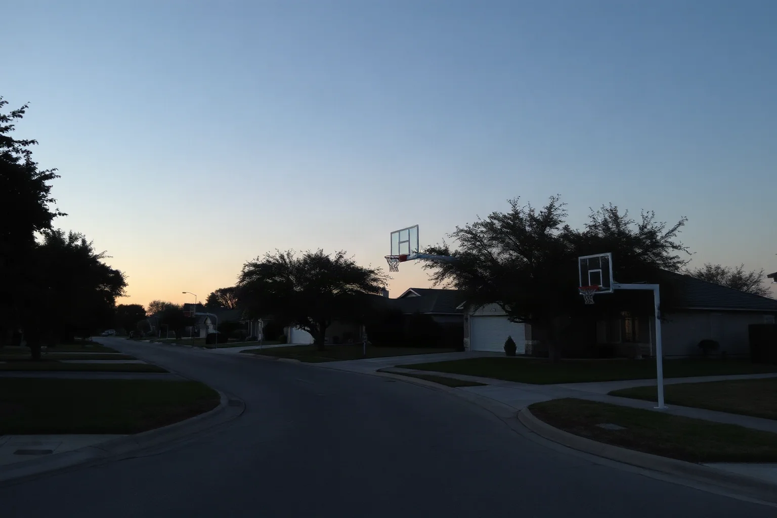 A suburban San Antonio street at dusk with basketball hoops visible in driveways, empty and quiet with long shadows