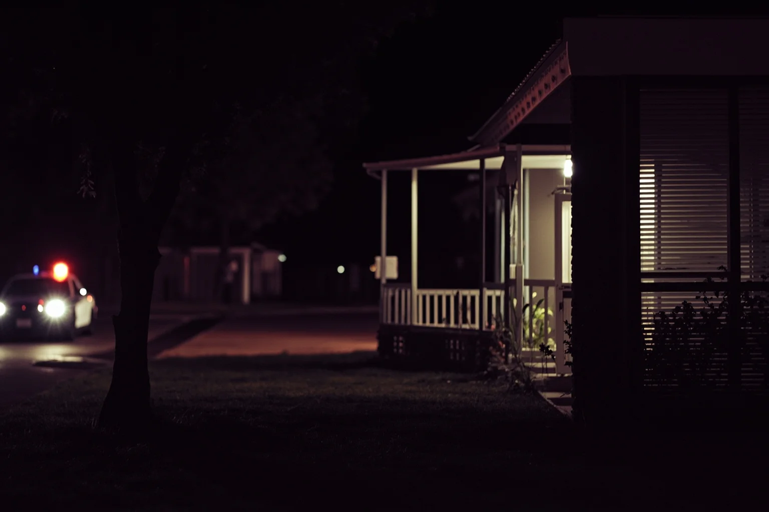 A vintage 1950s suburban house exterior at night with police car lights in the distance
