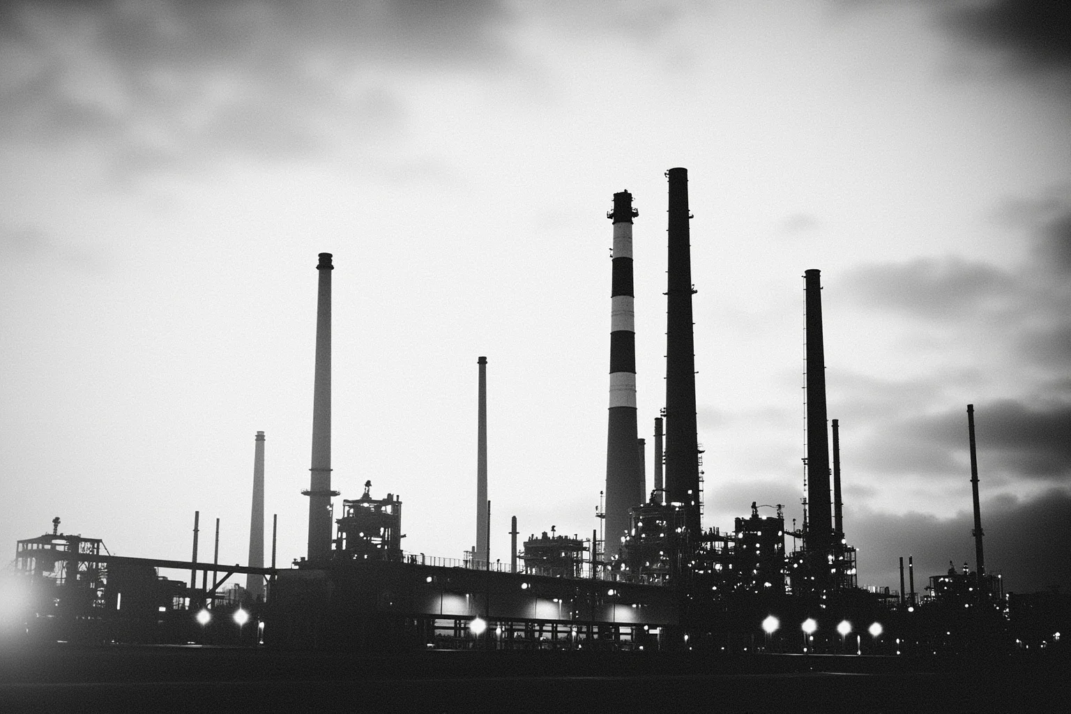 A vintage black-and-white style oil refinery with tall smokestacks against the sky
