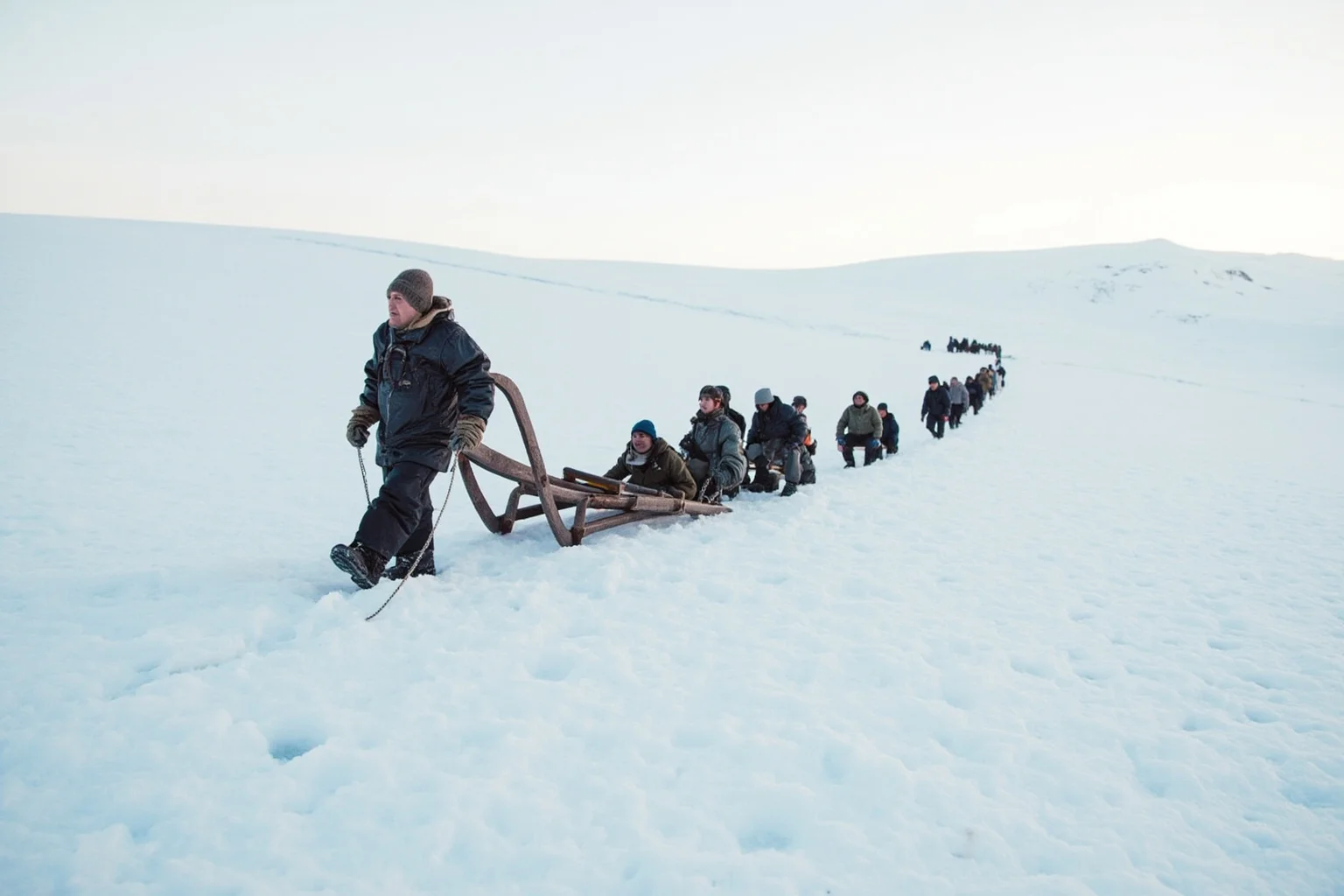 A weary explorer and small team hauling a wooden sledge across a vast snowy Antarctic plain under a pale sky