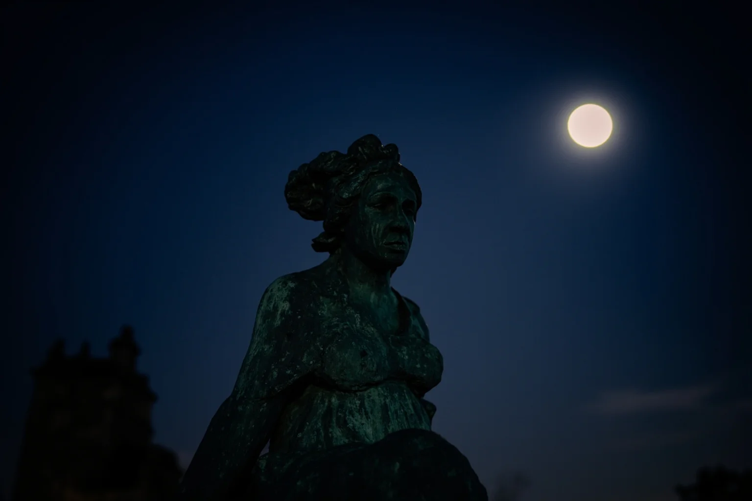 A weathered bronze cemetery statue of a mourning woman under moonlight