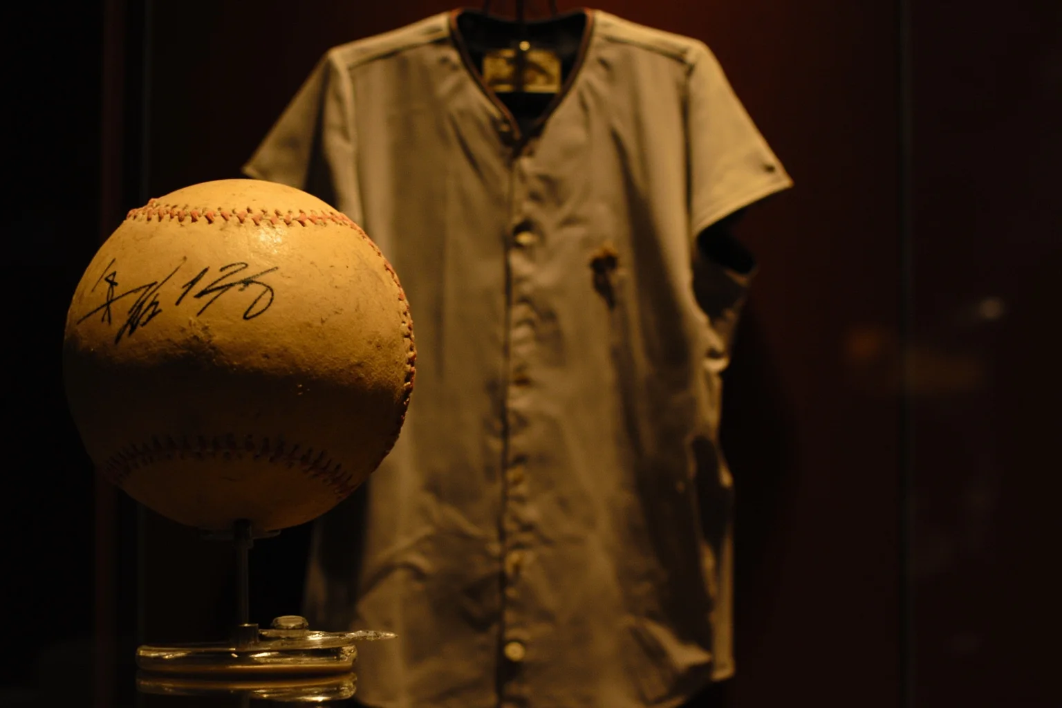 A weathered vintage baseball jersey and signed ball displayed in a glass memorabilia case, warm museum lighting