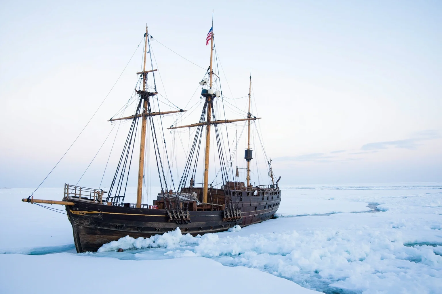 A wooden Antarctic-era sailing ship trapped in cracking pack ice under a pale polar sky