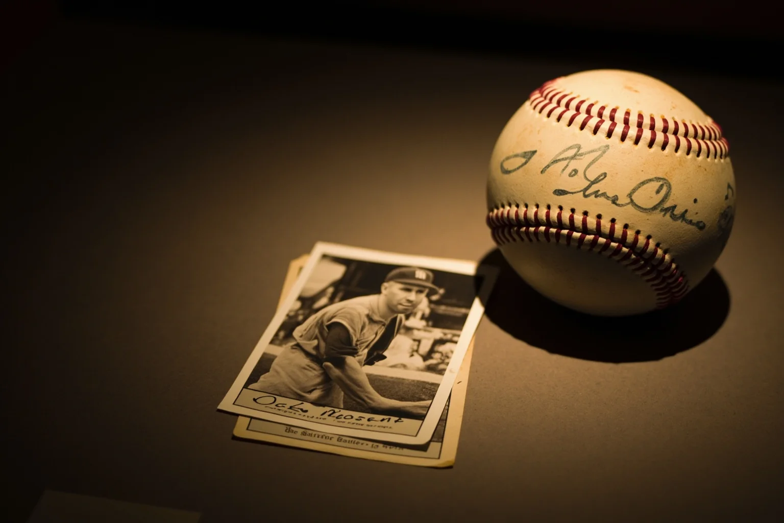 An old sepia-toned baseball card and a weathered vintage baseball with faded signature ink, soft museum lighting