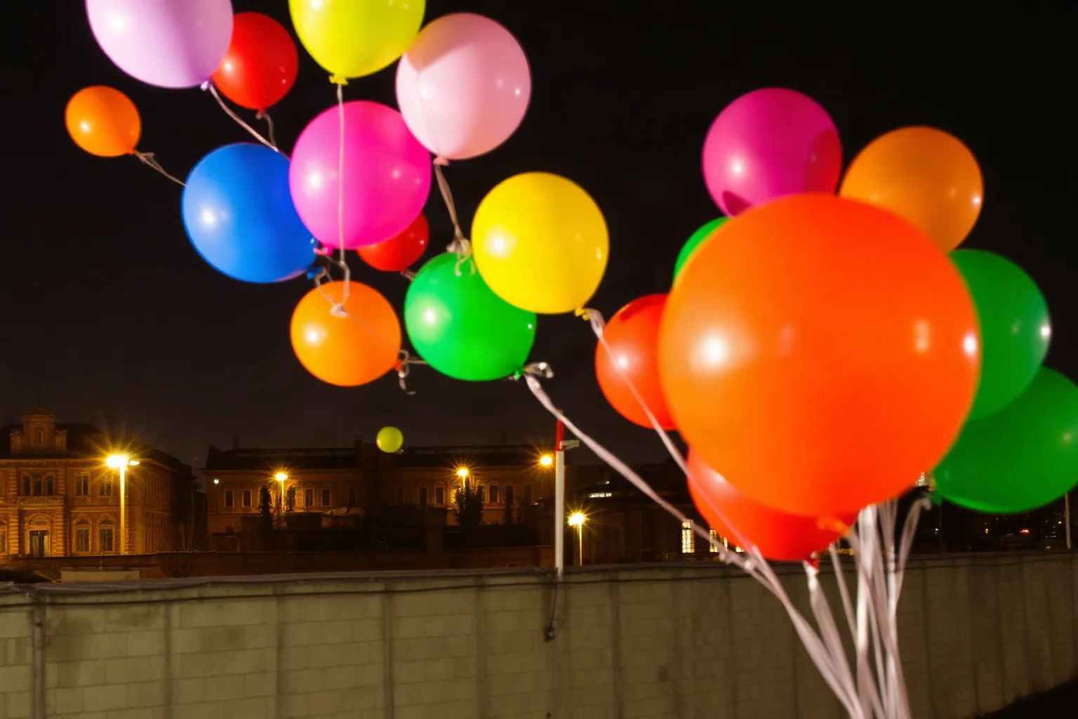 Colorful balloons drifting across a fortified wall dividing two sectors of a Cold War-era divided city at night