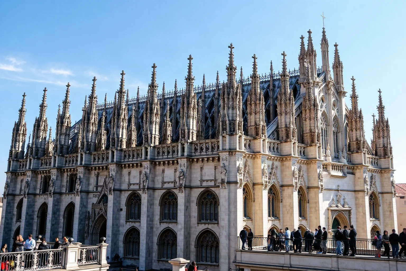 Enormous Gothic marble cathedral covered in spires and statues, people walking on the rooftop terrace, bright sky