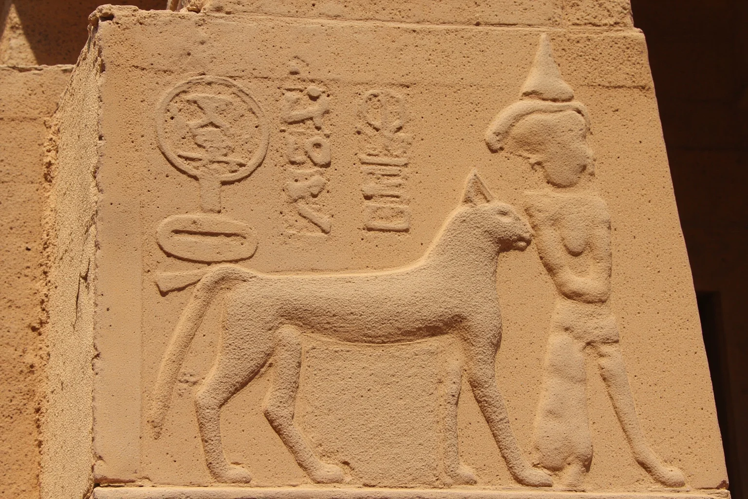 Ancient Egyptian stone relief carving of a sacred cat beside a goddess figure, sandy temple setting