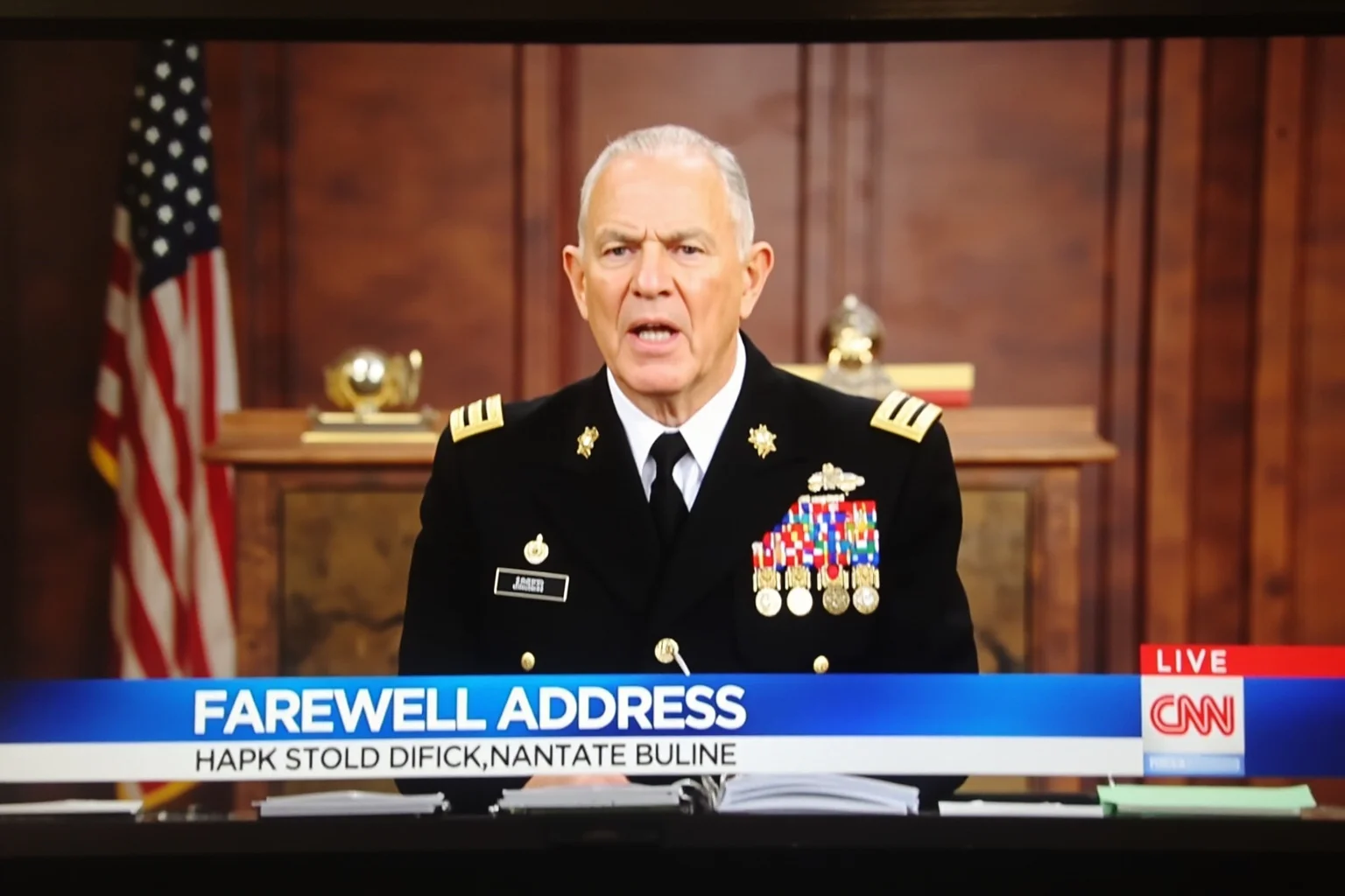Illustration of an older statesman in military-style attire giving a televised farewell address from a wood-paneled office