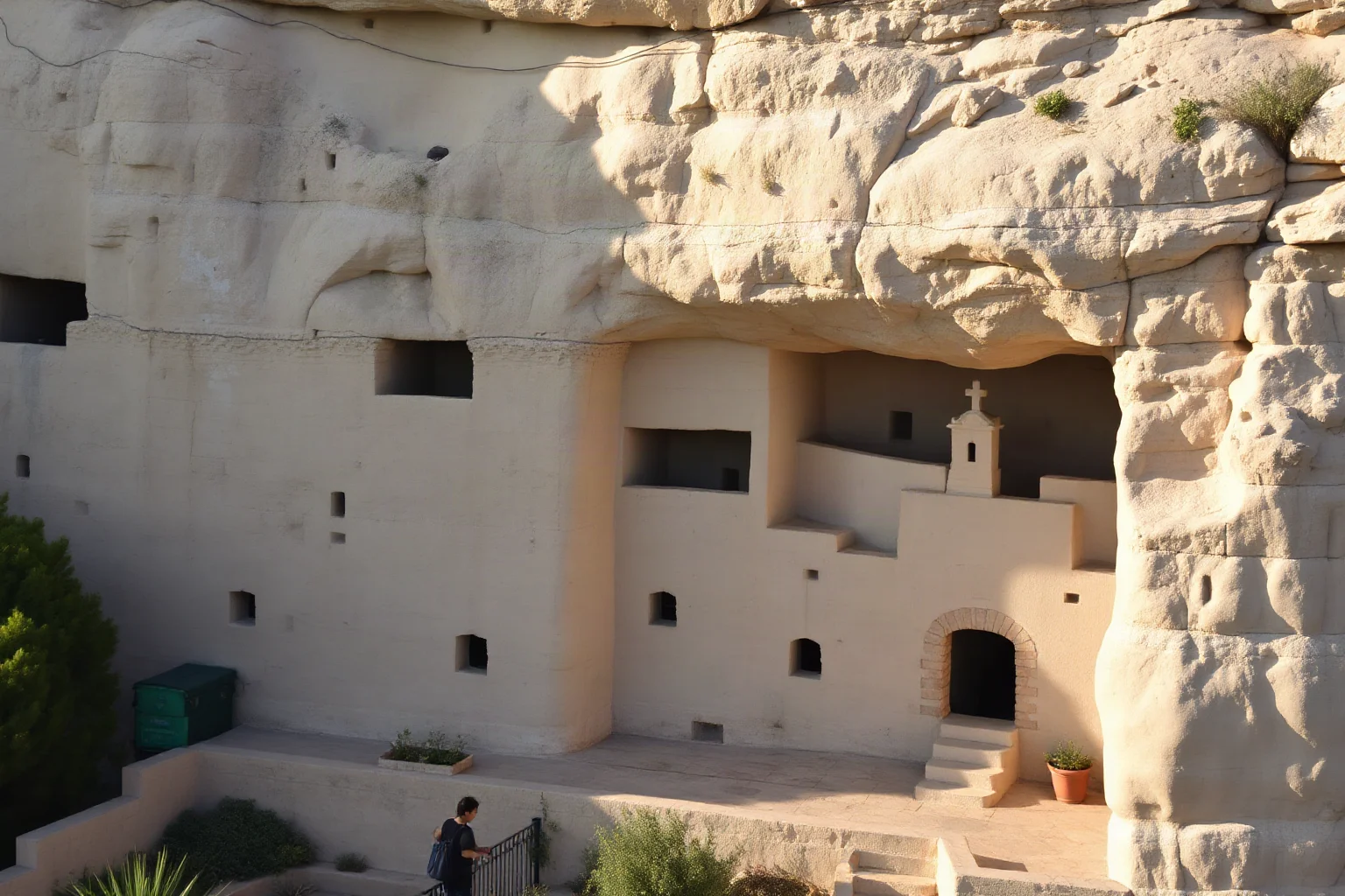Monastic cells carved into a pale rock cliff face with a small chapel entrance, Mediterranean hillside, soft morning light