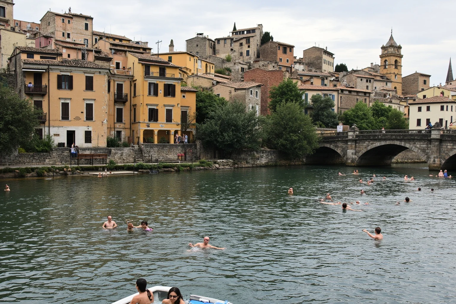 People swimming and relaxing in a river near a medieval town with a bathhouse in the background