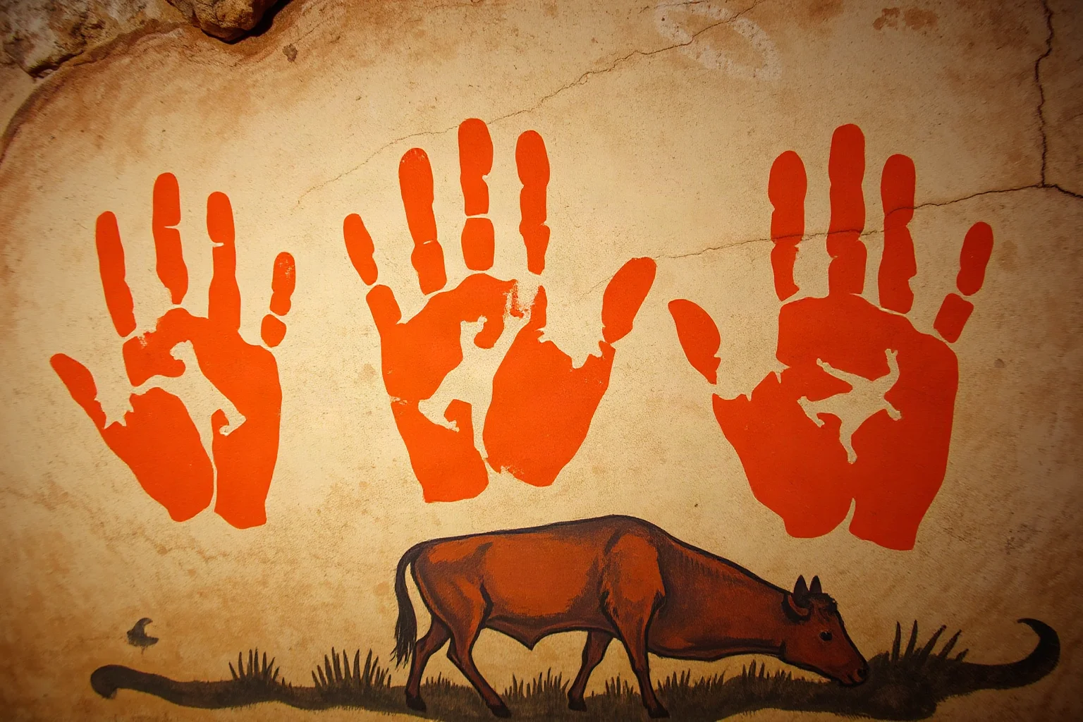 Reddish-orange hand stencils layered on a cave wall with an animal painting of a wild cow below, showing different time periods