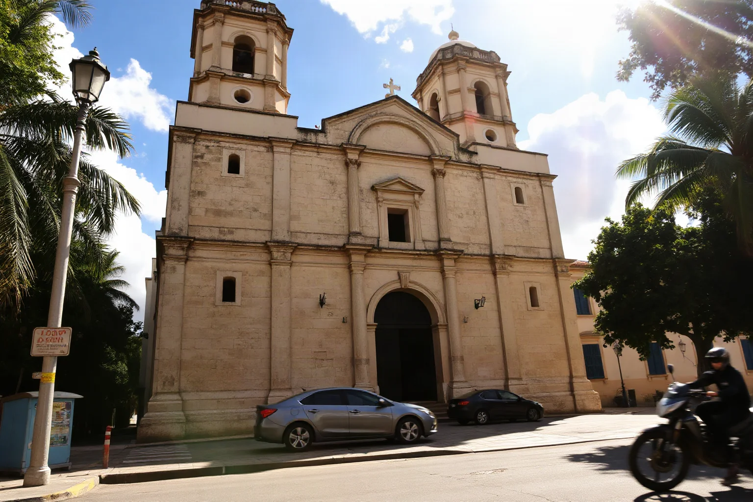 Exterior of a weathered stone Caribbean colonial cathedral with twin bell towers under bright tropical sunlight, old city street in foreground