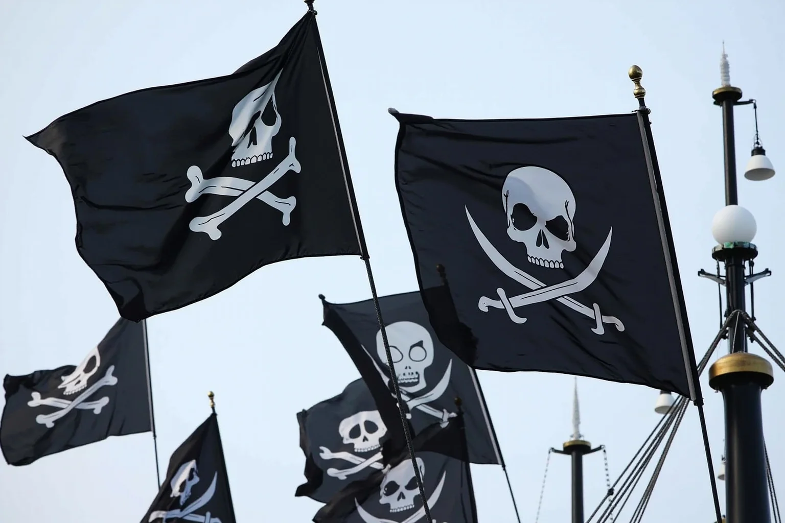 Several different pirate flags with varied skull, skeleton, and hourglass designs flying from ship masts