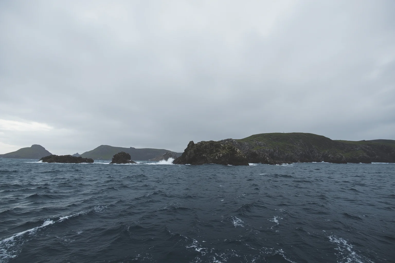 A rocky, windswept island archipelago in the North Atlantic seen from a boat, dramatic grey sky, no people