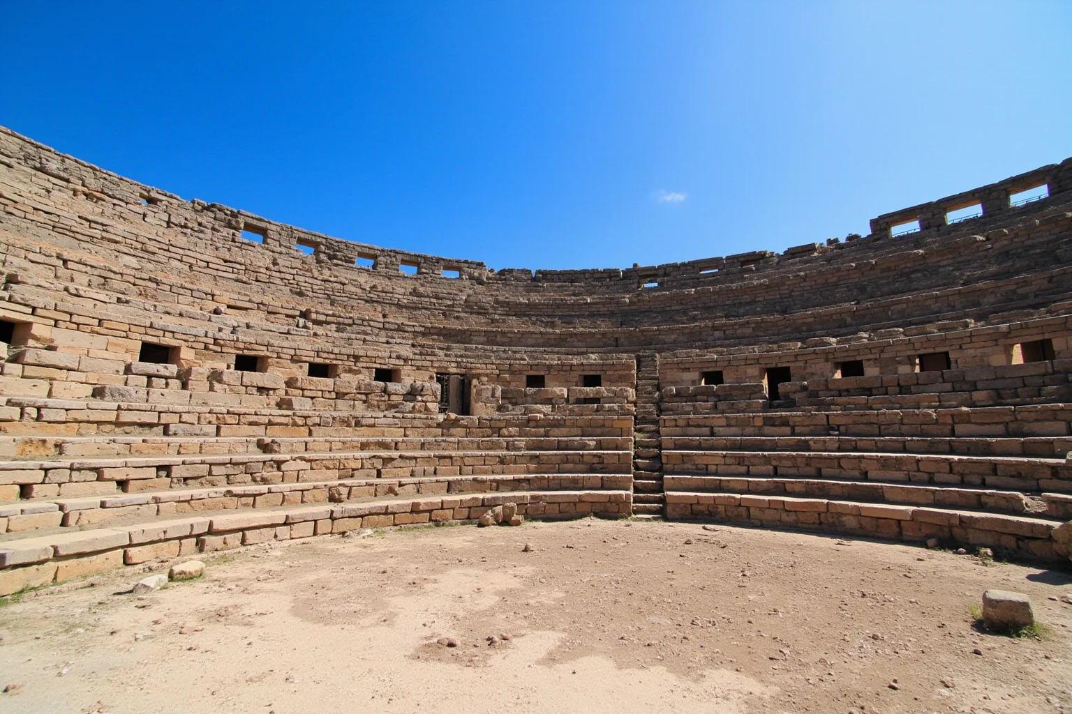 Wide shot of a weathered stone amphitheater ruin with tiered seating under a blue sky