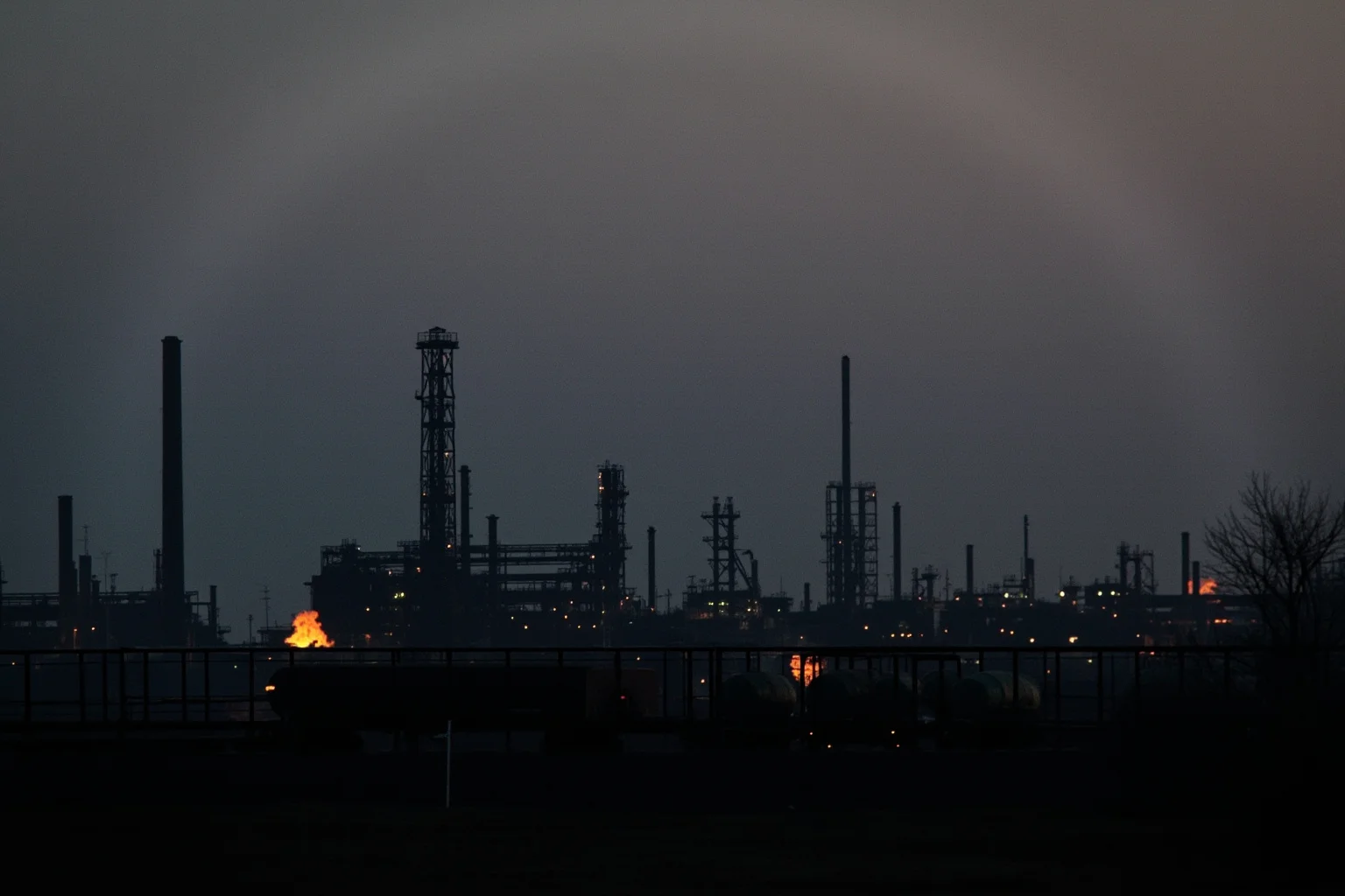 Old Money: 24 Hidden Dynasties That Shaped the Course of History 20 A 1900s oil refinery skyline at dusk with steel towers and rail tanker cars lit by gas flares, no people, moody industrial landscape photography.