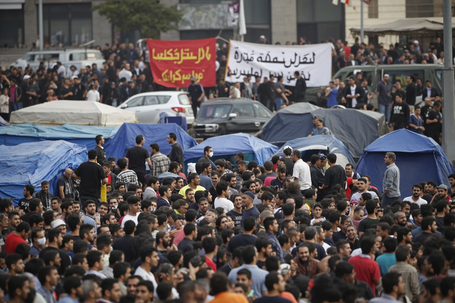22 Evil Dictators Who Were Eventually Overthrown 20 A crowded Tahrir Square with tents and Arabic banners, police in the background, blurred figures.