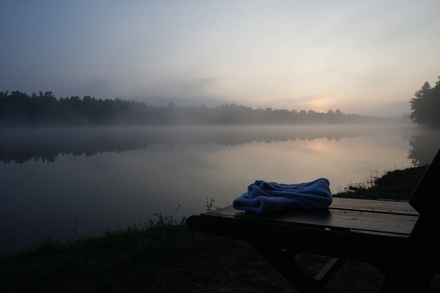 A misty riverbank at sunrise with a folded towel and wooden bench on the shore, no people.
