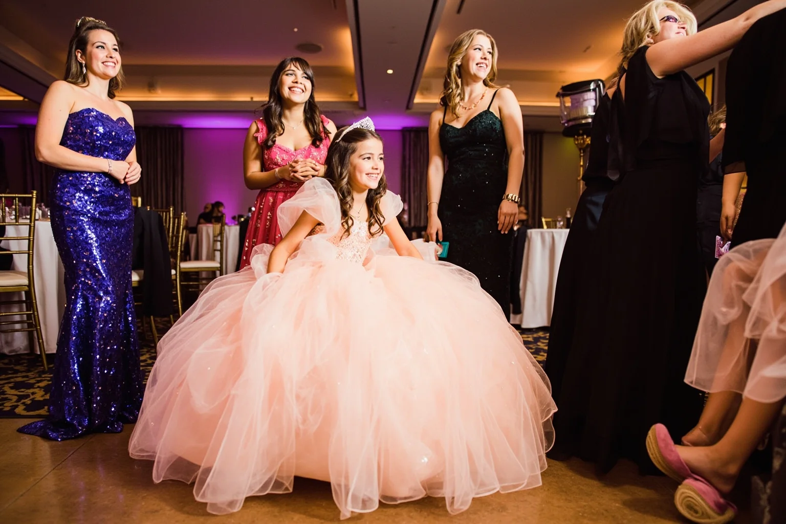 A smiling girl in a big pastel quinceañera gown surrounded by her court of honor in formal attire at a festive ballroom