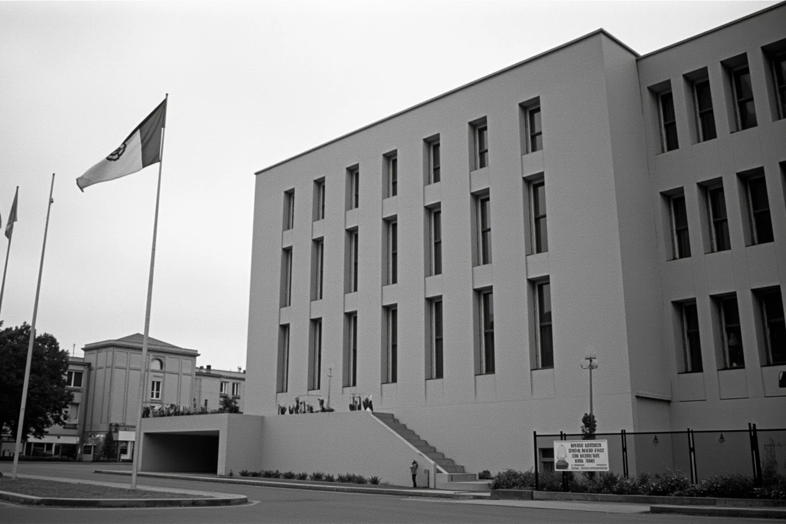 A United Nations building exterior with flags at half-mast, overcast sky, somber documentary photography style, 1960s era