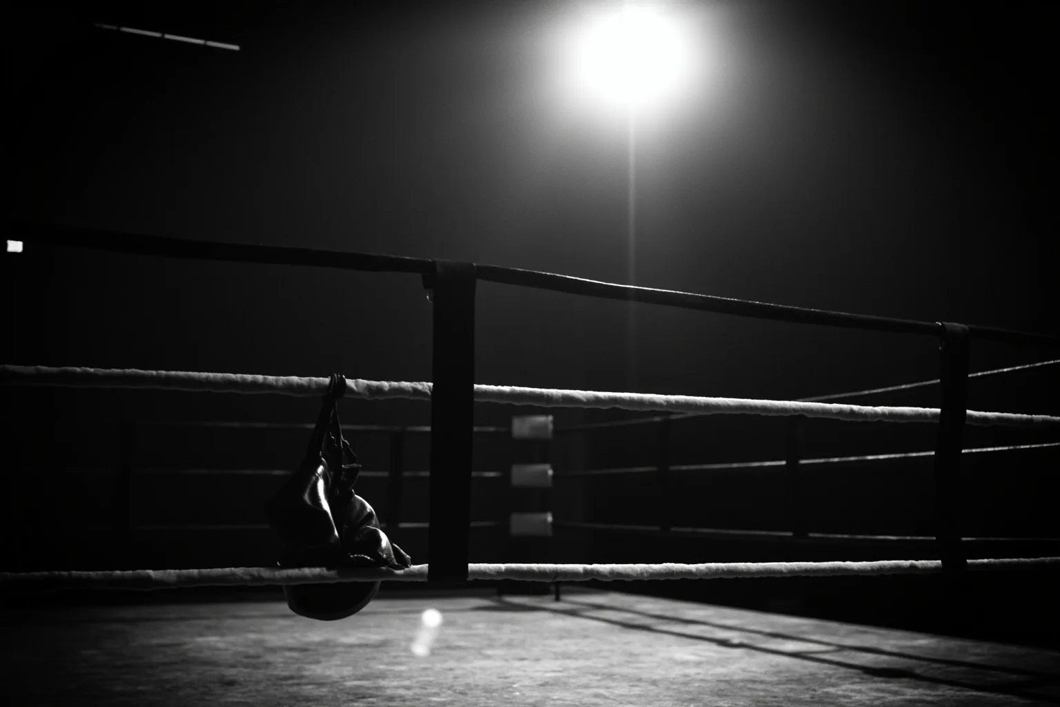 A vintage 1960s boxing ring with empty gloves hanging on the ropes, dramatic spotlight, black and white photography style